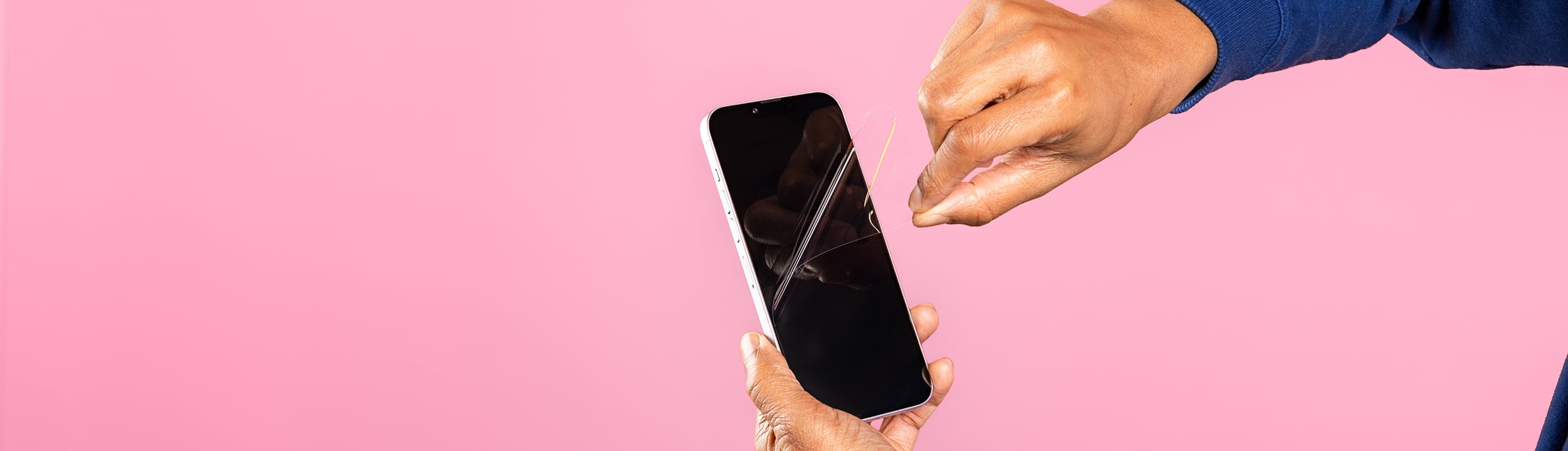 Zwei Hände entfernen eine Folien-Schutzfolie von einem iPhone, der Hintergrund ist rosa.