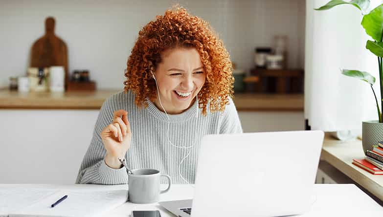 Eine Frau sitzt vor ihrem Laptop, lacht auf den Bildschirm und trägt Ohrhörer mit Kabeln.