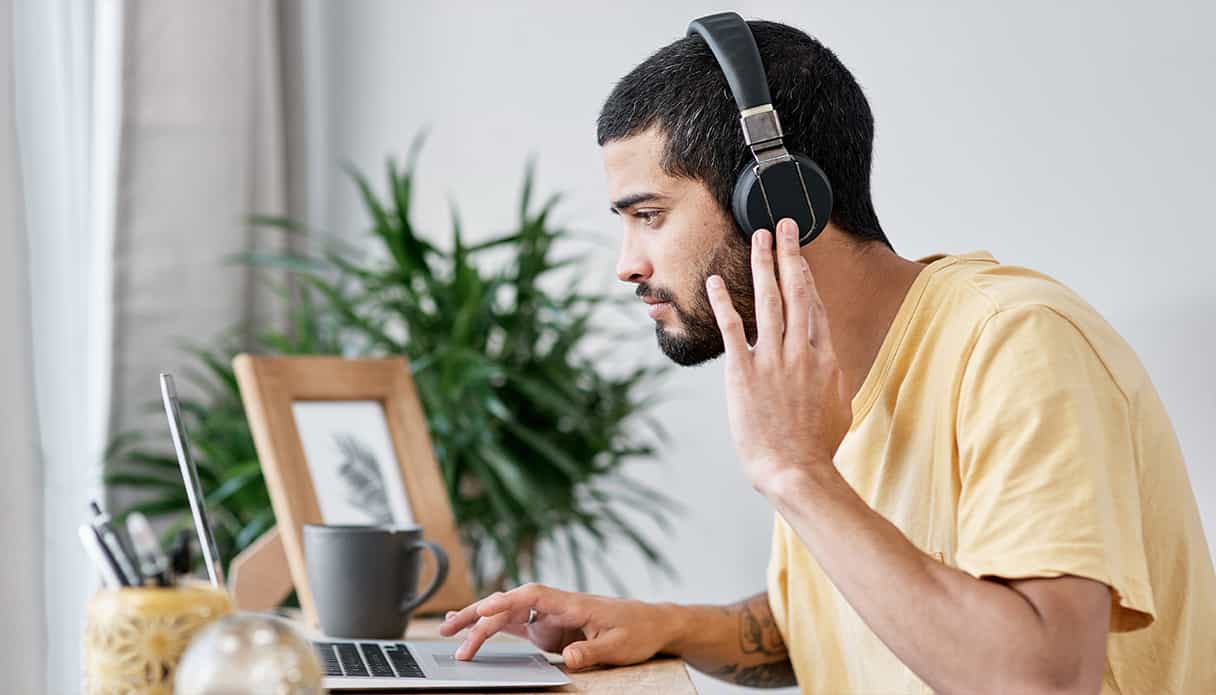 Ein Mann sitzt am Schreibtisch und schaut auf seinen Laptop, während er geräuschunterdrückende Kopfhörer trägt.