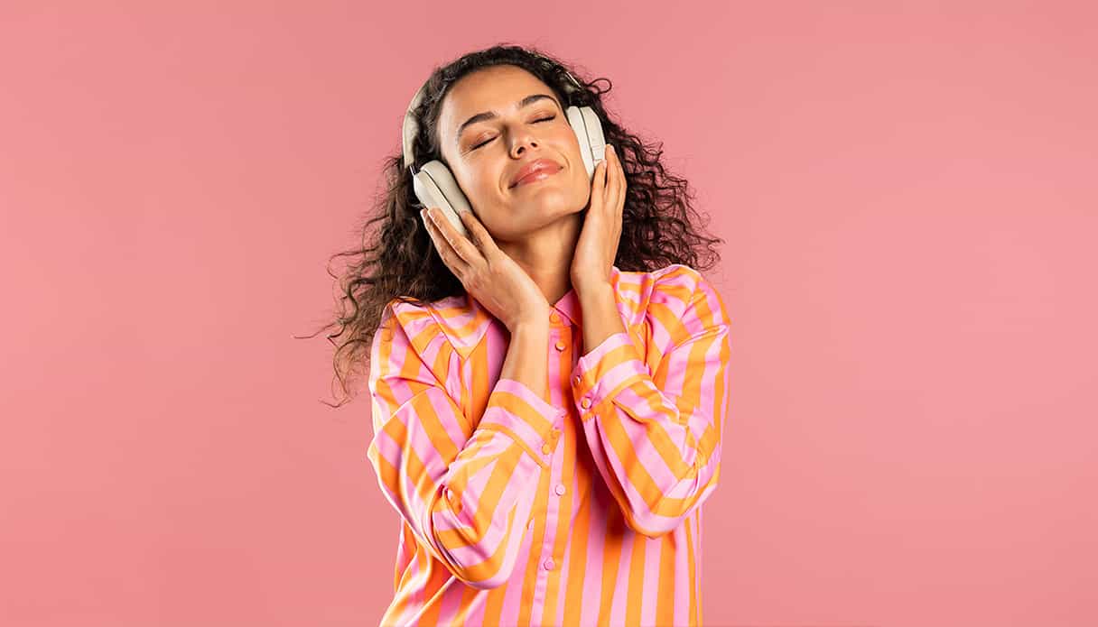 Eine Frau auf einem rosa Hintergrund genießt Musik mit geschlossenen Augen durch ihre geräuschunterdrückenden Kopfhörer.