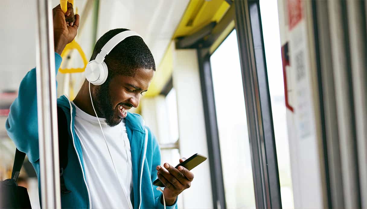 Ein Mann steht im Bus und schaut lachend auf sein Telefon, er trägt einen kabelgebundenen Kopfhörer.