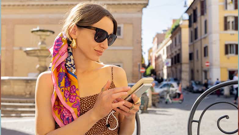 Eine Frau mit Sonnenbrille sitzt auf einer Terrasse und benutzt ihr Telefon und eine Powerbank.