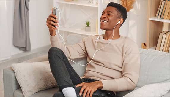 Ein Mann sitzt auf der Couch, videotelefoniert über sein Telefon und trägt Ohrhörer mit Kabel.