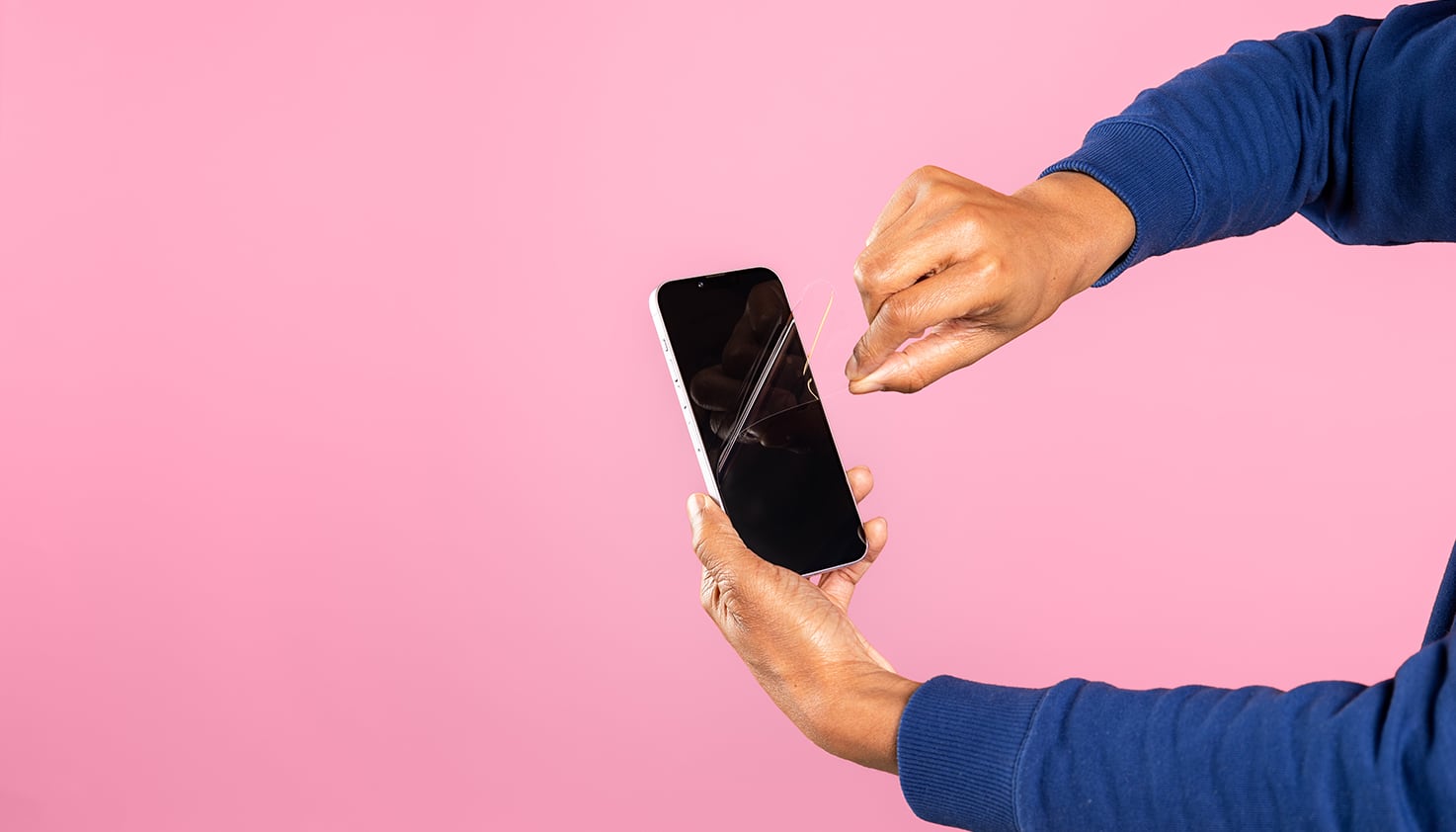 Zwei Hände entfernen eine Folien-Schutzfolie von einem iPhone, der Hintergrund ist rosa.