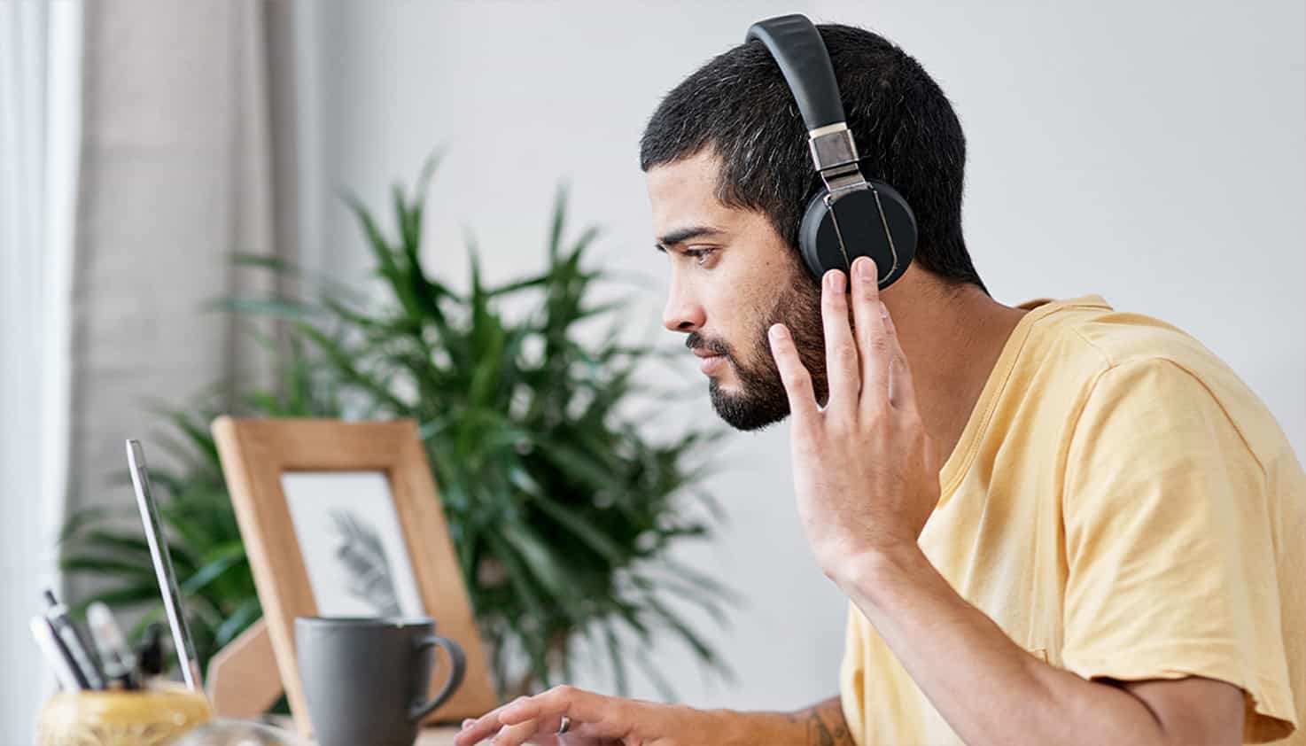 Ein Mann sitzt am Schreibtisch und schaut auf seinen Laptop, während er geräuschunterdrückende Kopfhörer trägt.