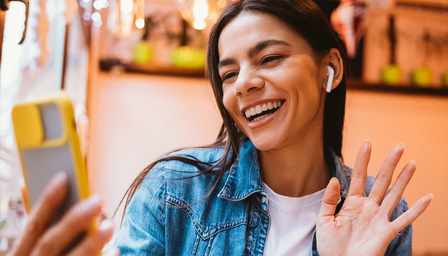 Eine Frau winkt lächelnd ihrem Telefon zu, sie ist am Videotelefonieren und trägt ihre kabellosen Ohrhörer.