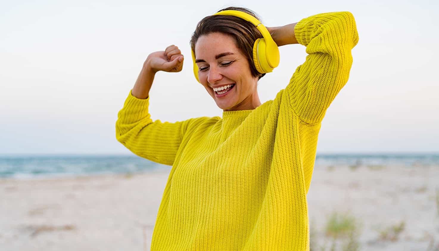 Am Strand trägt eine lachende Frau einen hellgelben Lärm reduzierenden Kopfhörer auf dem Kopf.